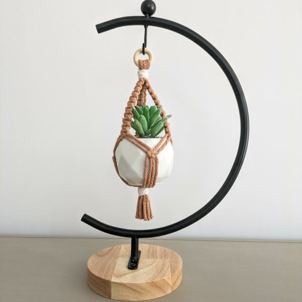 Mini macrame plant hanger with a small white pot and artificial green succulent included. SHown hanging on a black metal half moon display stand with a natural wood base. Plant hanger is shown in salmon pink cord.
