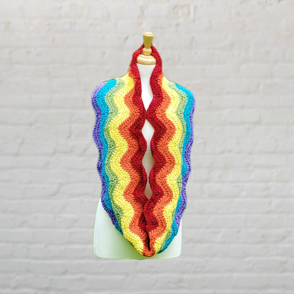 A crochet infinity scarf made with wavy stripes in all the colors of the rainbow. It is looped around the neck of a dress form on a blurred white brick background.
